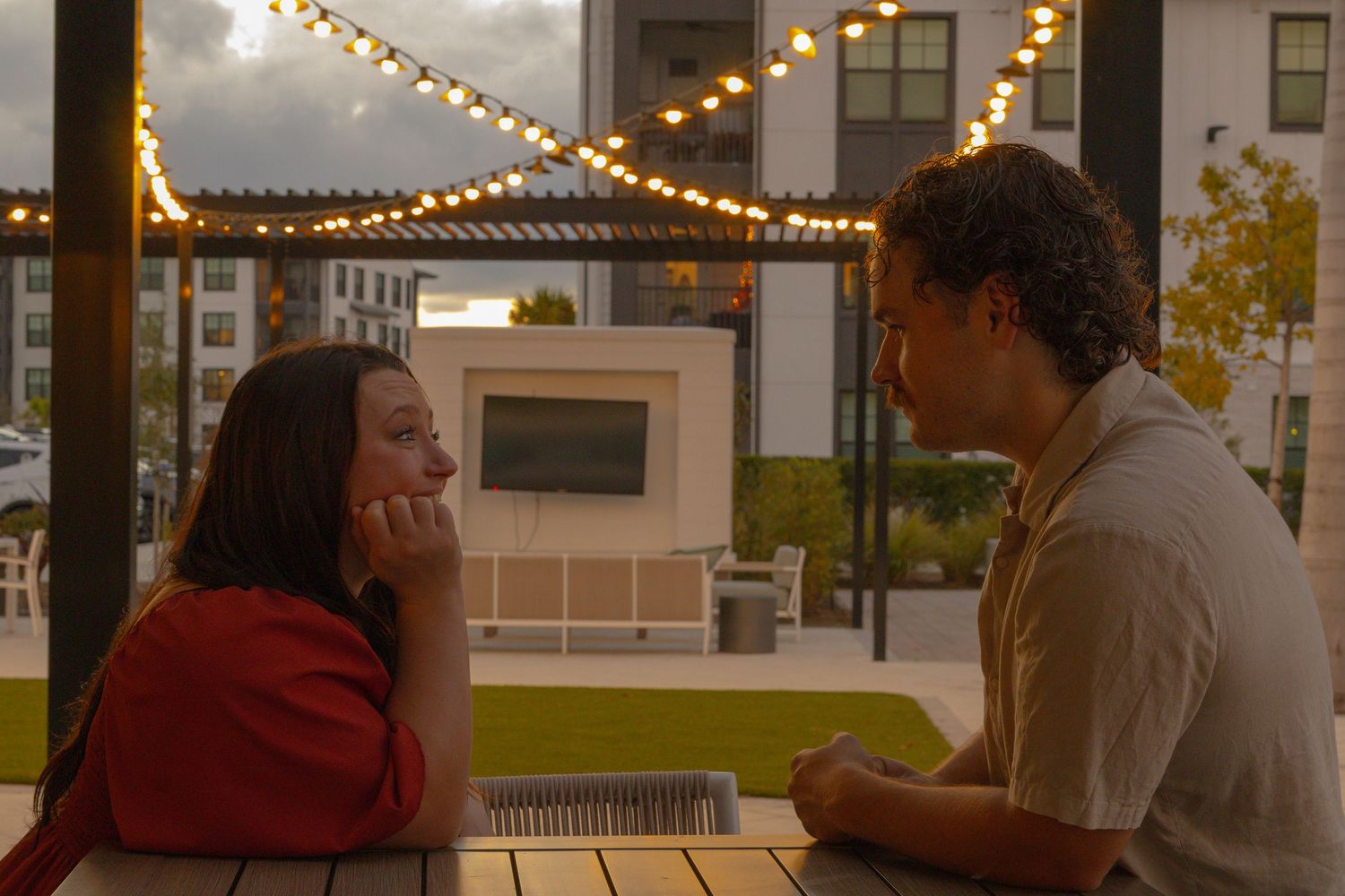 Two people share an intimate conversation at an outdoor patio table under string lights at dusk.