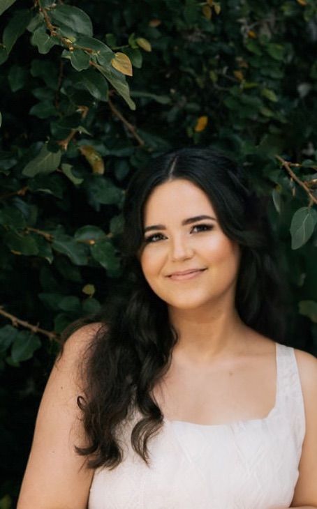 Young woman with dark wavy hair smiling outdoors in a white dress, framed by lush green foliage.