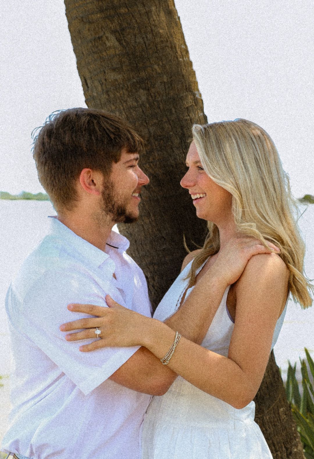 A smiling couple hugs near a tree by the water during an outdoor engagement photo session.