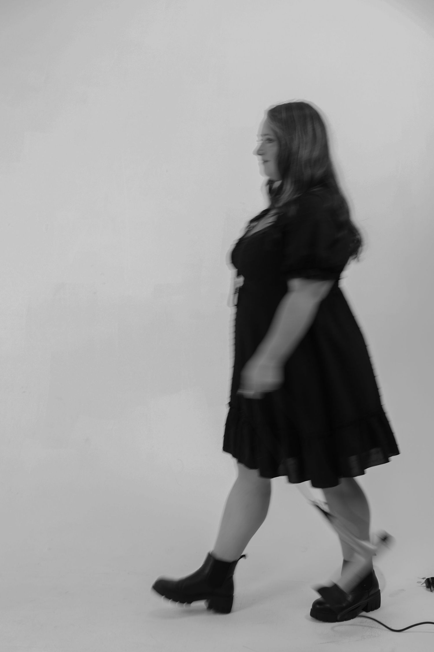 Black and white motion blur photo of a woman in a black dress walking across a white background.
