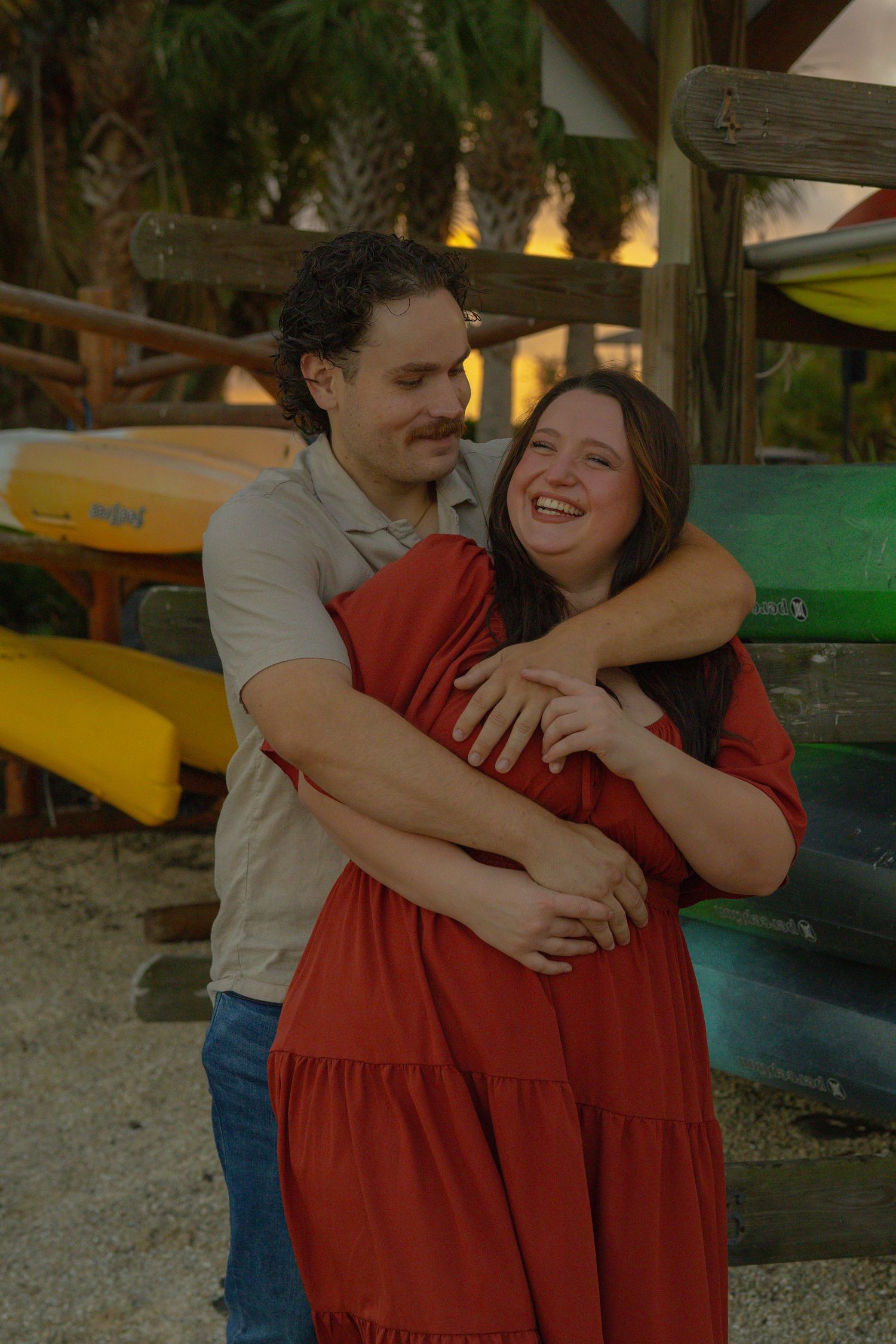 Happy couple laughing and embracing in front of colorful kayaks at sunset.