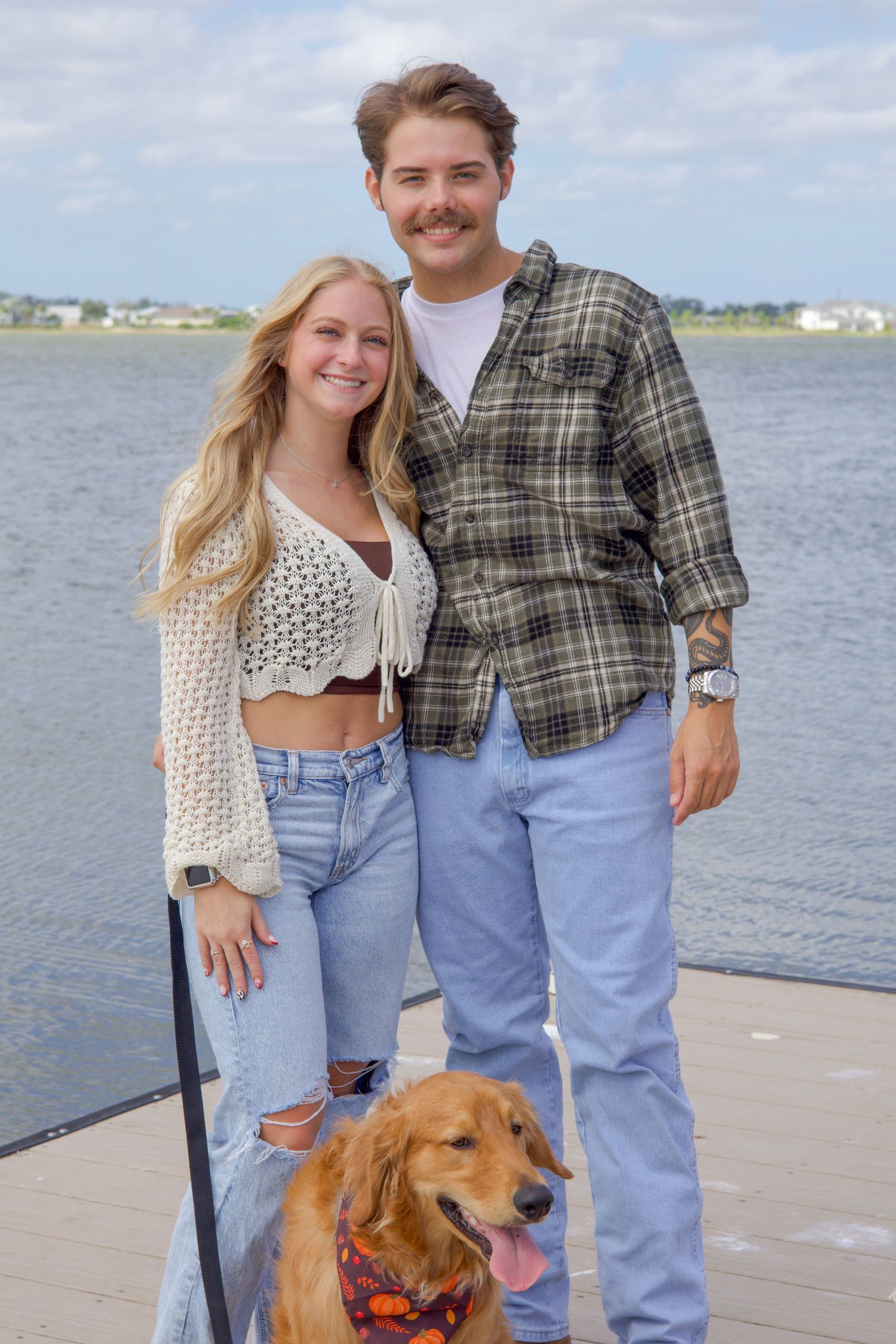 Smiling couple standing on dock with golden retriever wearing bandana by water.