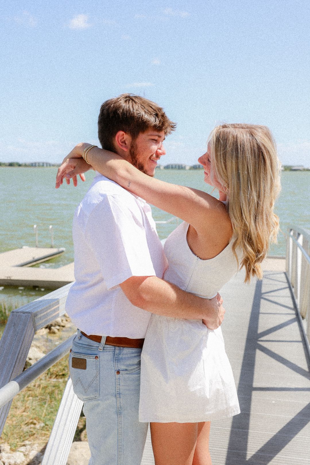 A smiling couple embraces on a sunny waterfront dock, wearing matching white outfits with blue sky behind them.