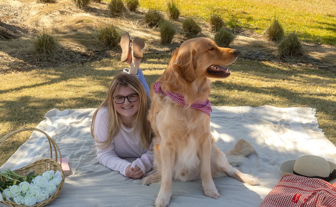 Girl lying on a picnic blanket smiling beside her Golden Retriever dog outdoors.