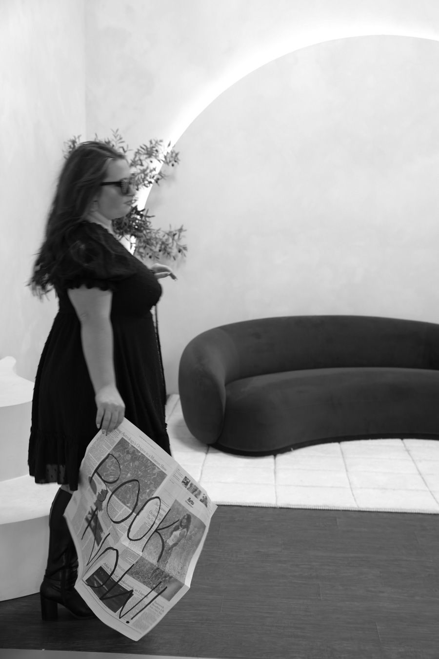 Woman in black dress holding a newspaper in a modern white interior space.