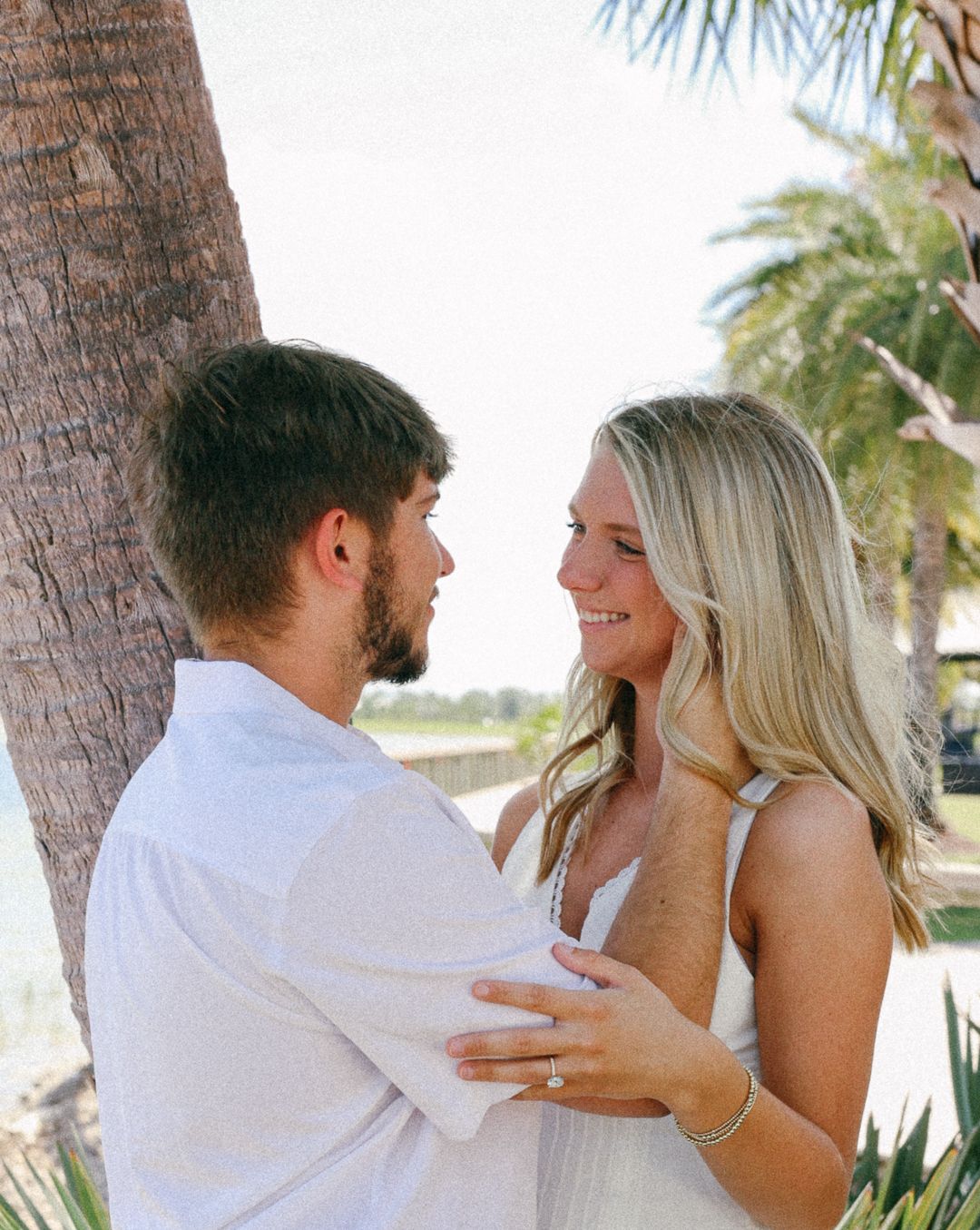 A happy couple holds each other close near a palm tree, with the woman displaying an engagement ring.