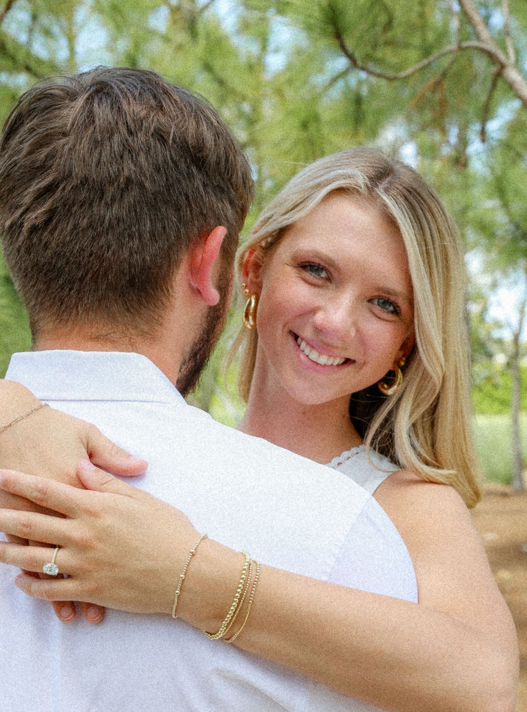 A smiling blonde woman in a white dress embraces her partner outdoors during an engagement photo session.