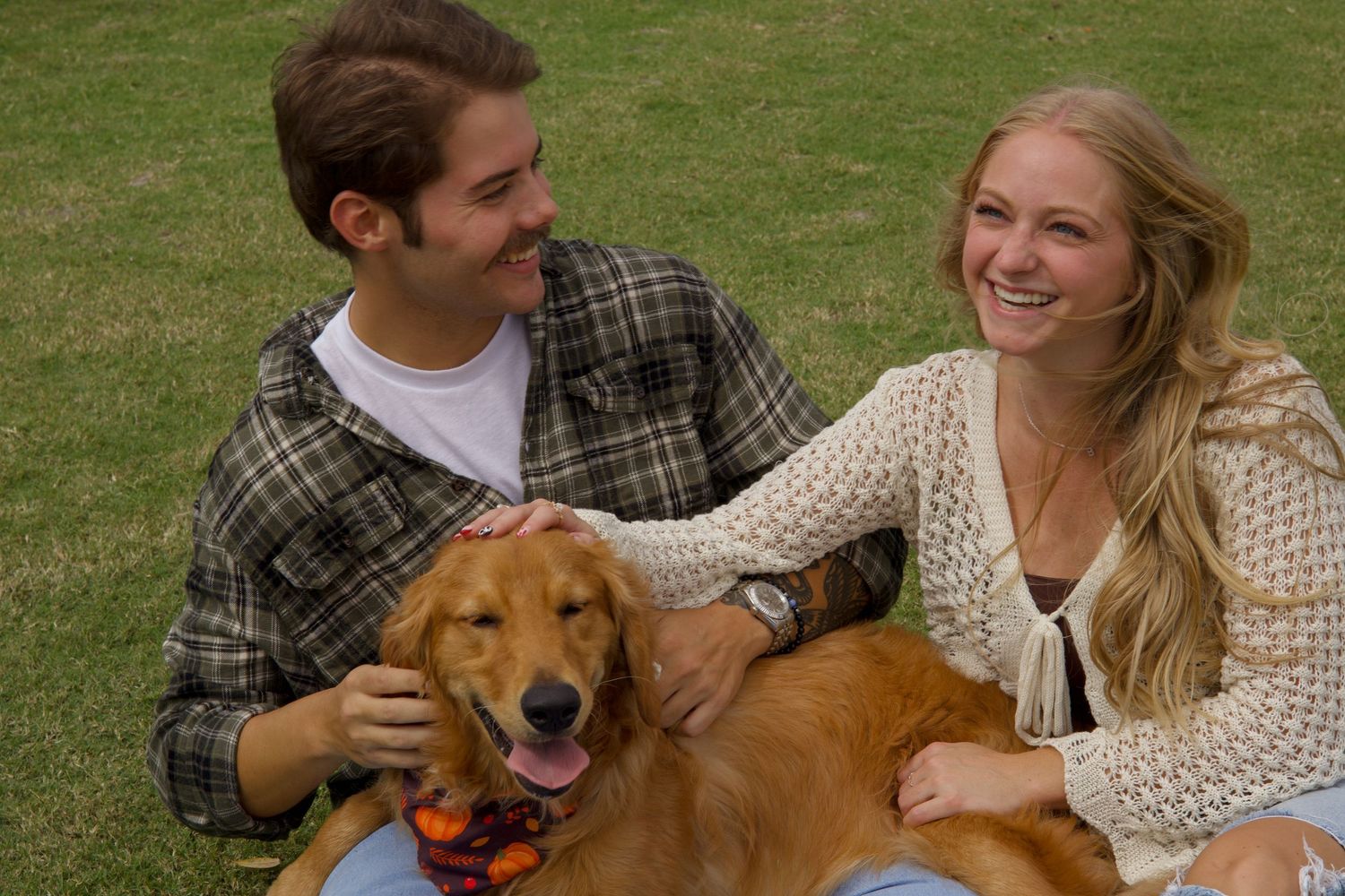 A young couple smiles while petting a happy golden retriever dog on a green grass lawn.