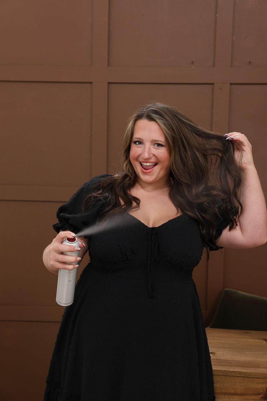 Woman in black dress laughing while spraying hairspray on her long brunette hair indoors.