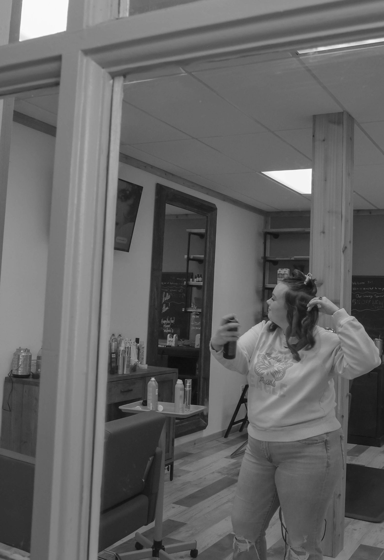 Black and white photo of a woman styling her hair with spray in a professional hair salon.