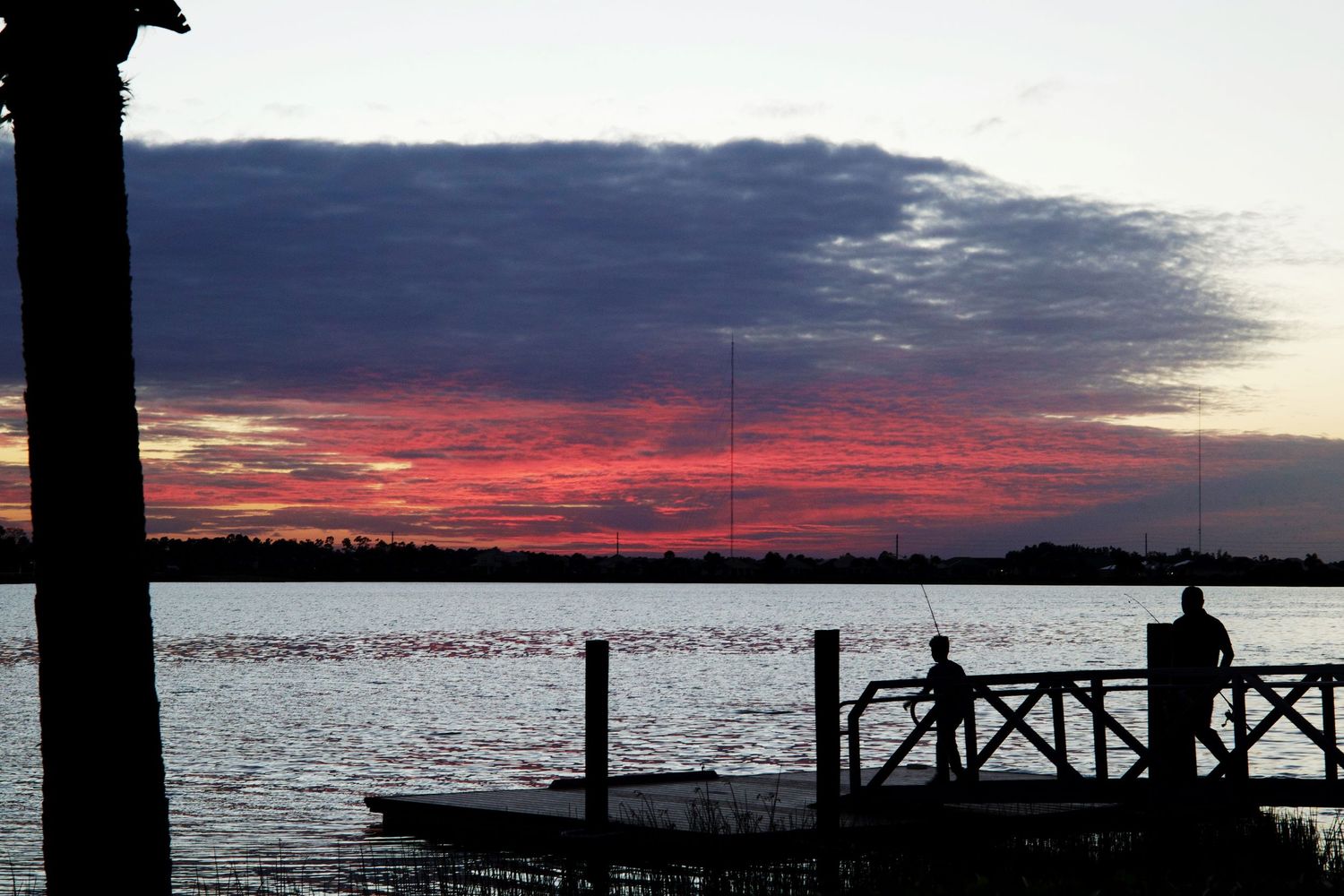 Silhouettes of people fishing on a dock at sunset over a calm lake with vibrant pink and purple skies.
