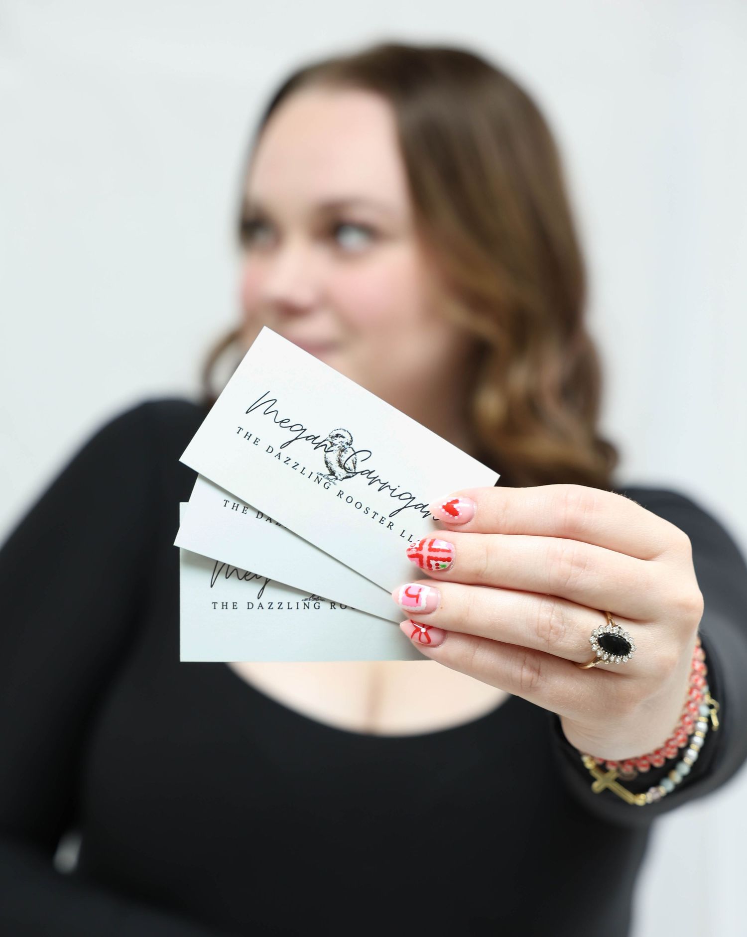 Woman holding multiple Megan Corrigan Dazzling Rooster LLC business cards with red manicured nails.