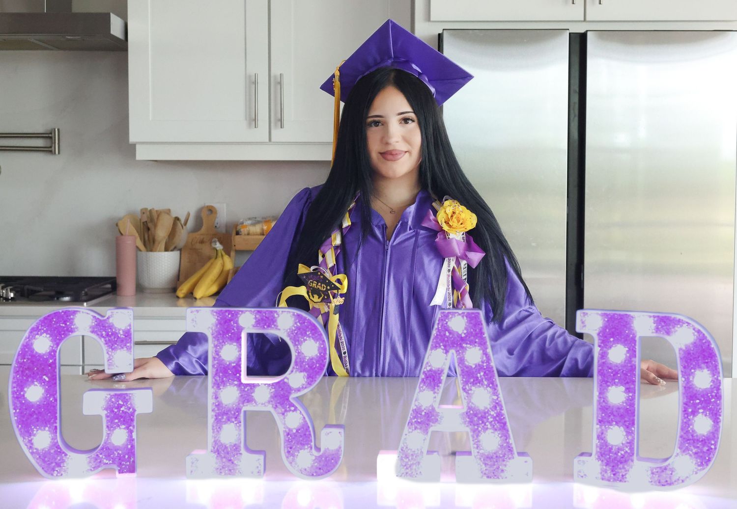 Young woman in purple graduation cap and gown posing behind illuminated GRAD letter signs in kitchen.