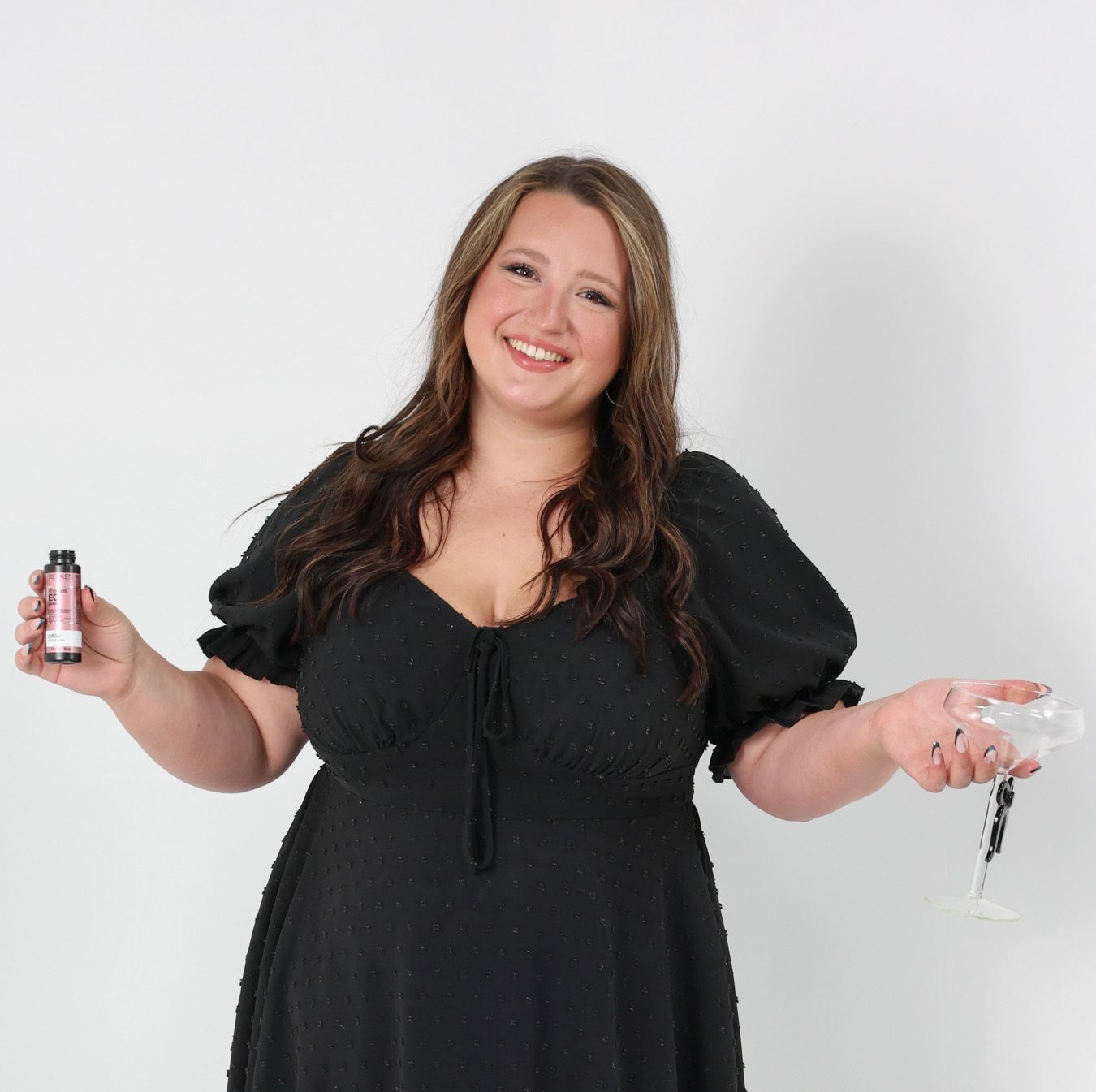 Smiling woman in black dress holding a small bottle and empty cocktail glass against white background.