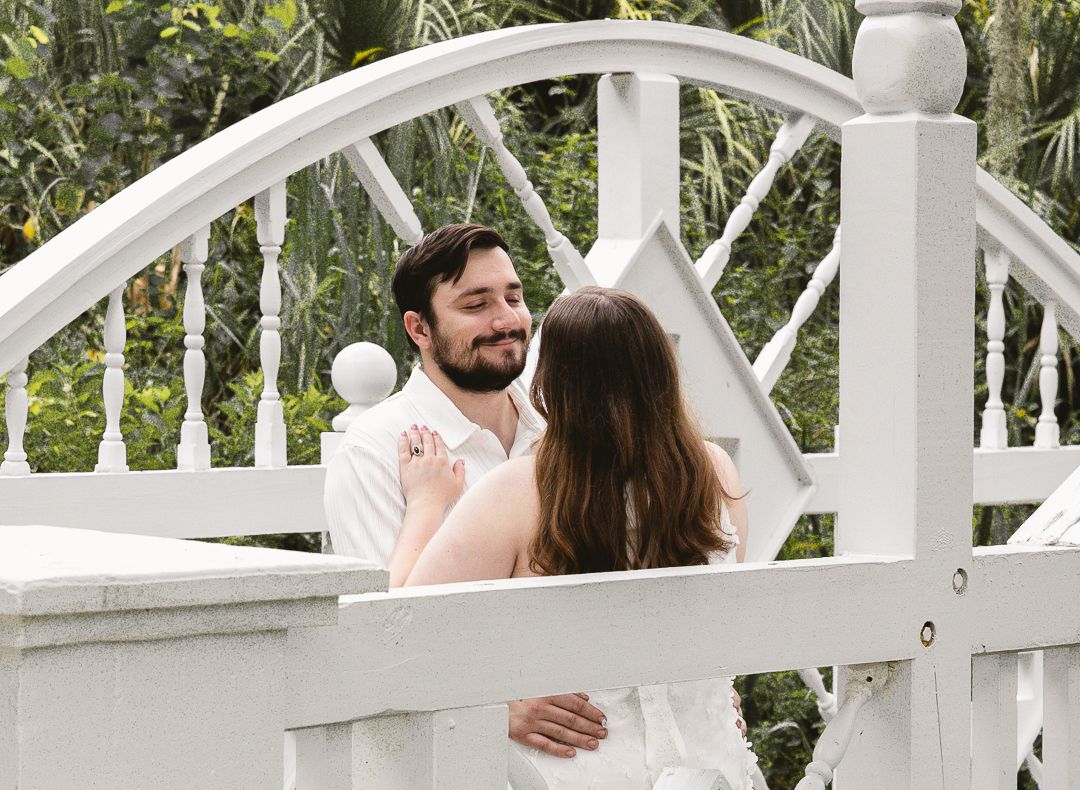 Couple embracing on a white bridge surrounded by lush greenery in a romantic outdoor setting.