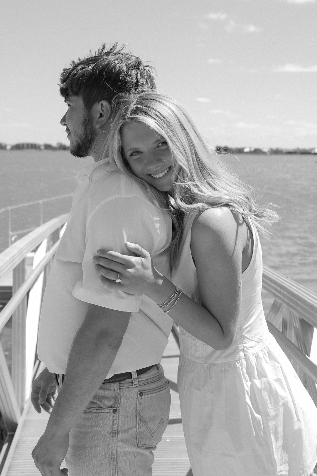 Black and white photo of smiling blonde woman hugging her fiance from behind on sunny waterfront dock.
