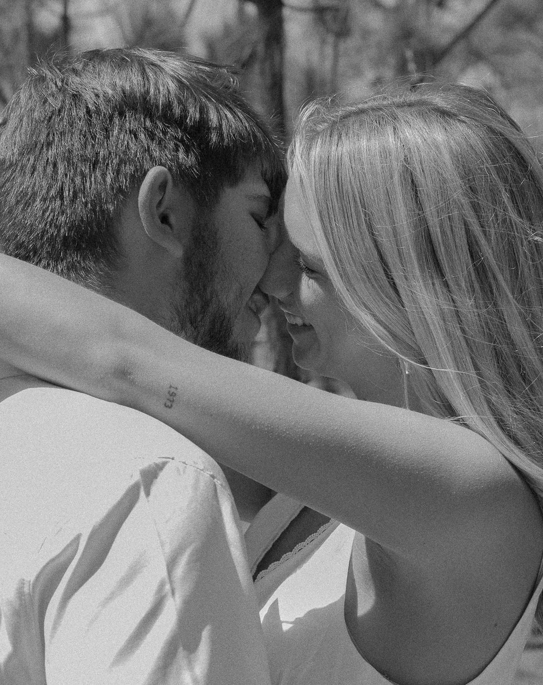 Black and white photo of couple touching foreheads and smiling intimately outdoors.