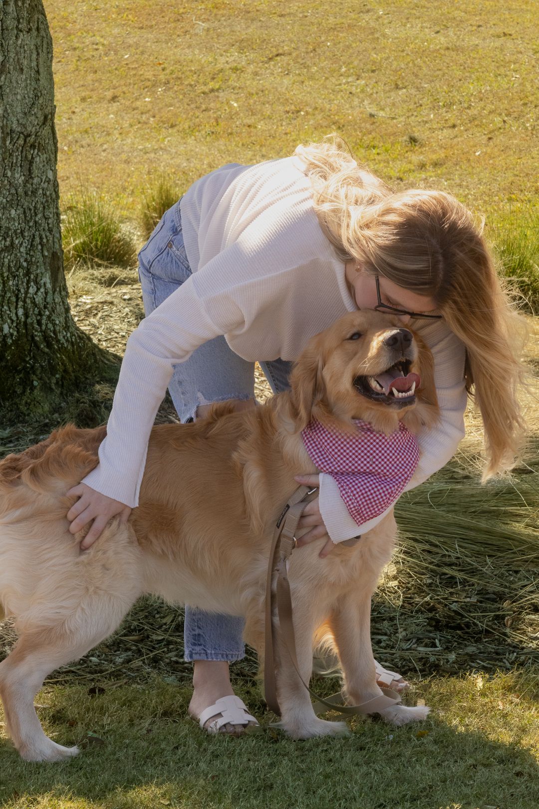 Blonde woman in white top leaning down to hug a golden retriever wearing a pink bandana.