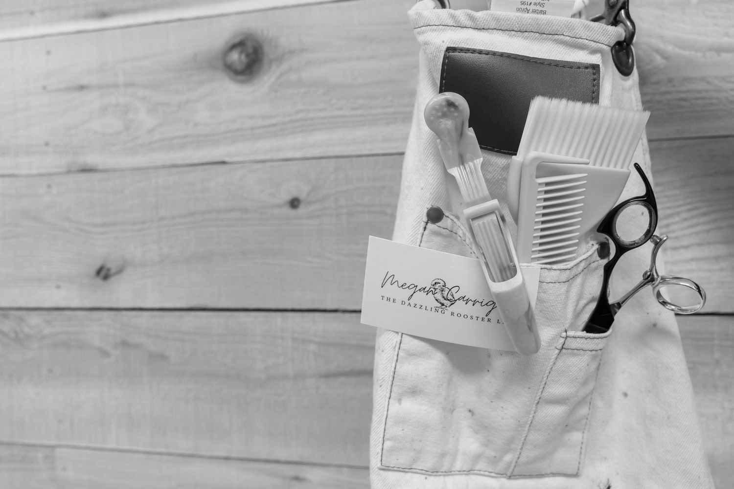 Black and white image of hair stylist apron with combs, scissors, and Megan Corrigan business card on wood background.