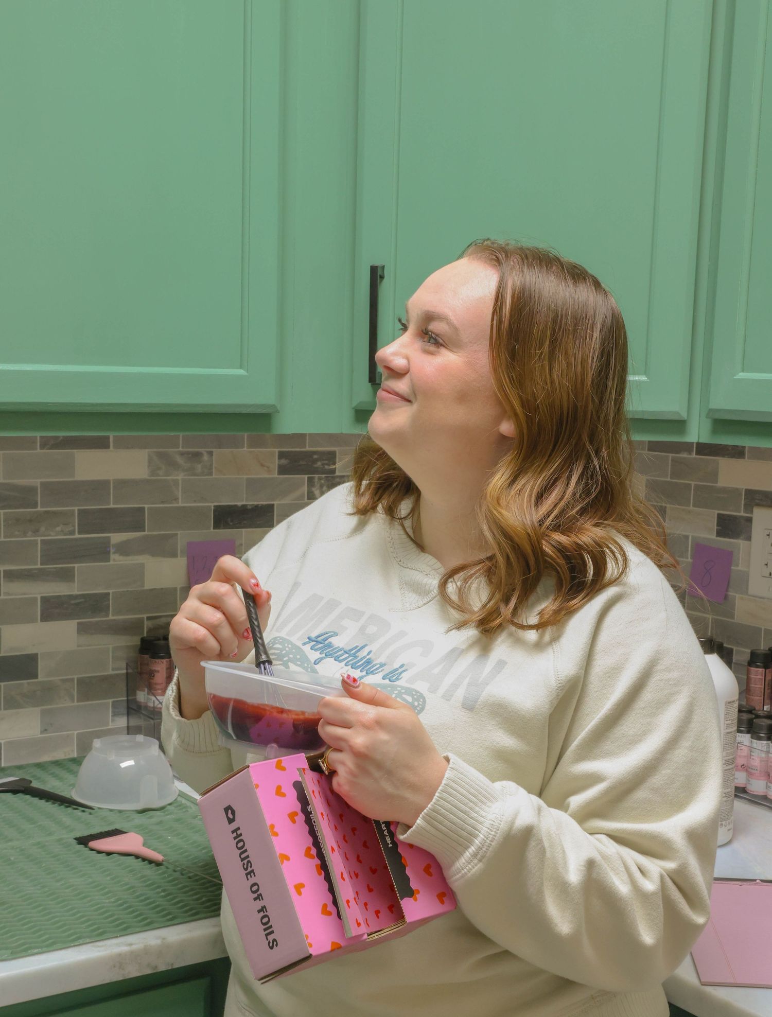 Smiling woman in cream sweatshirt holding a pink bag and tools near green cabinets in a salon.