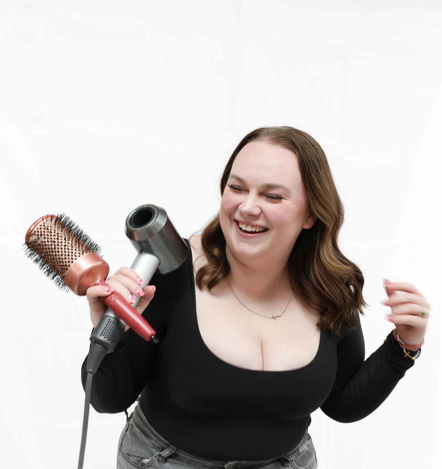 Smiling woman holding a Dyson hair dryer and round brush, wearing a black top on white background.