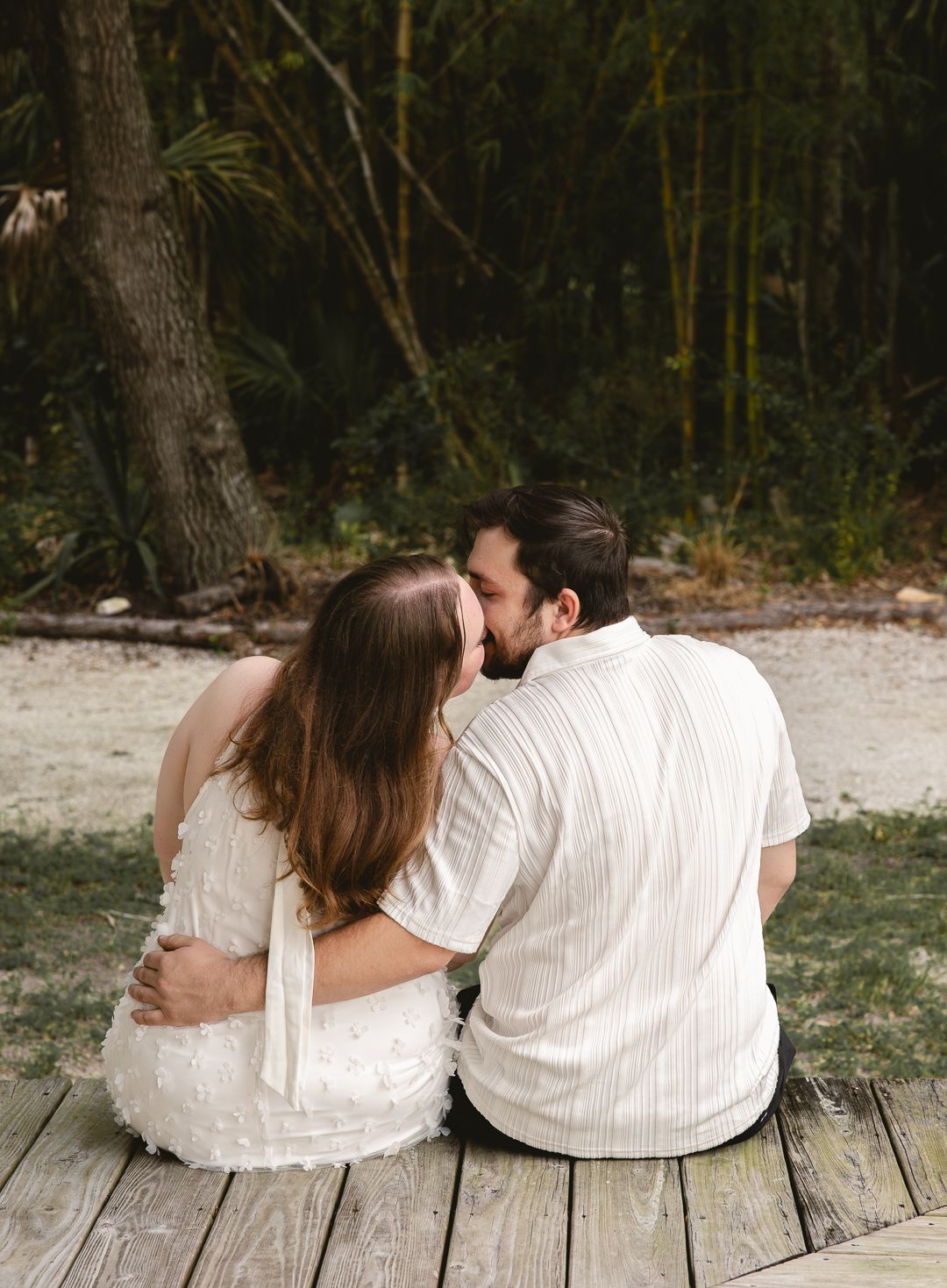 Couple sitting on wooden deck embracing and kissing with lush green trees in background.