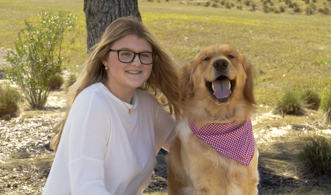 A smiling young woman poses with her golden retriever wearing a pink bandana in an outdoor field.