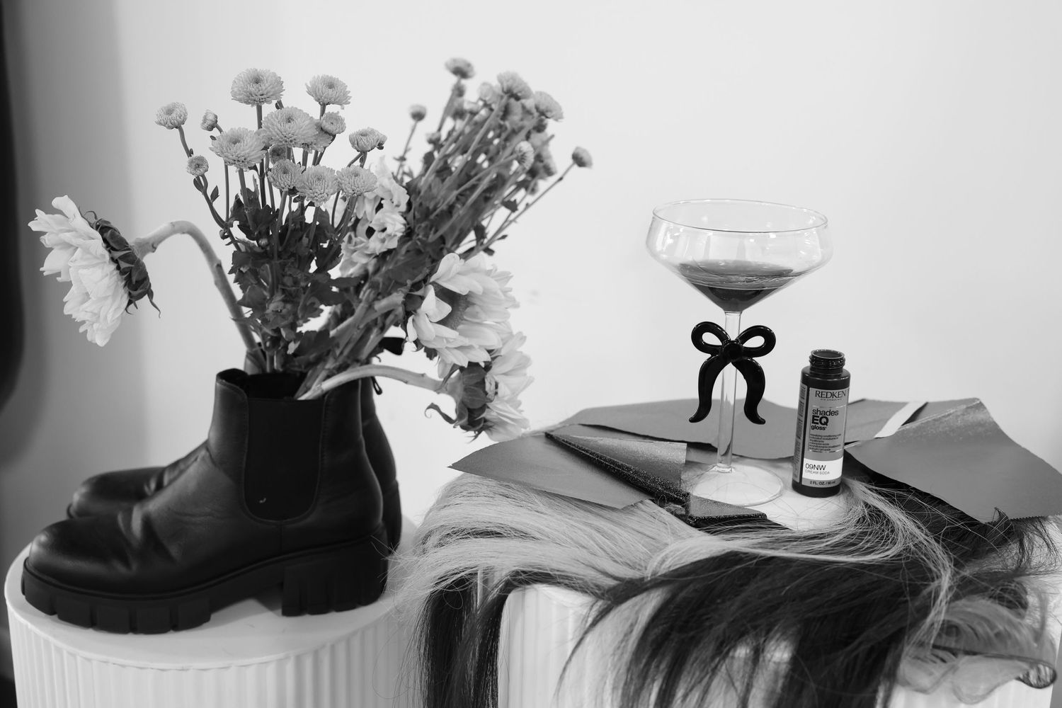 Black and white flat lay of boot vase with flowers, coupe glass, hair extensions, and bottle.