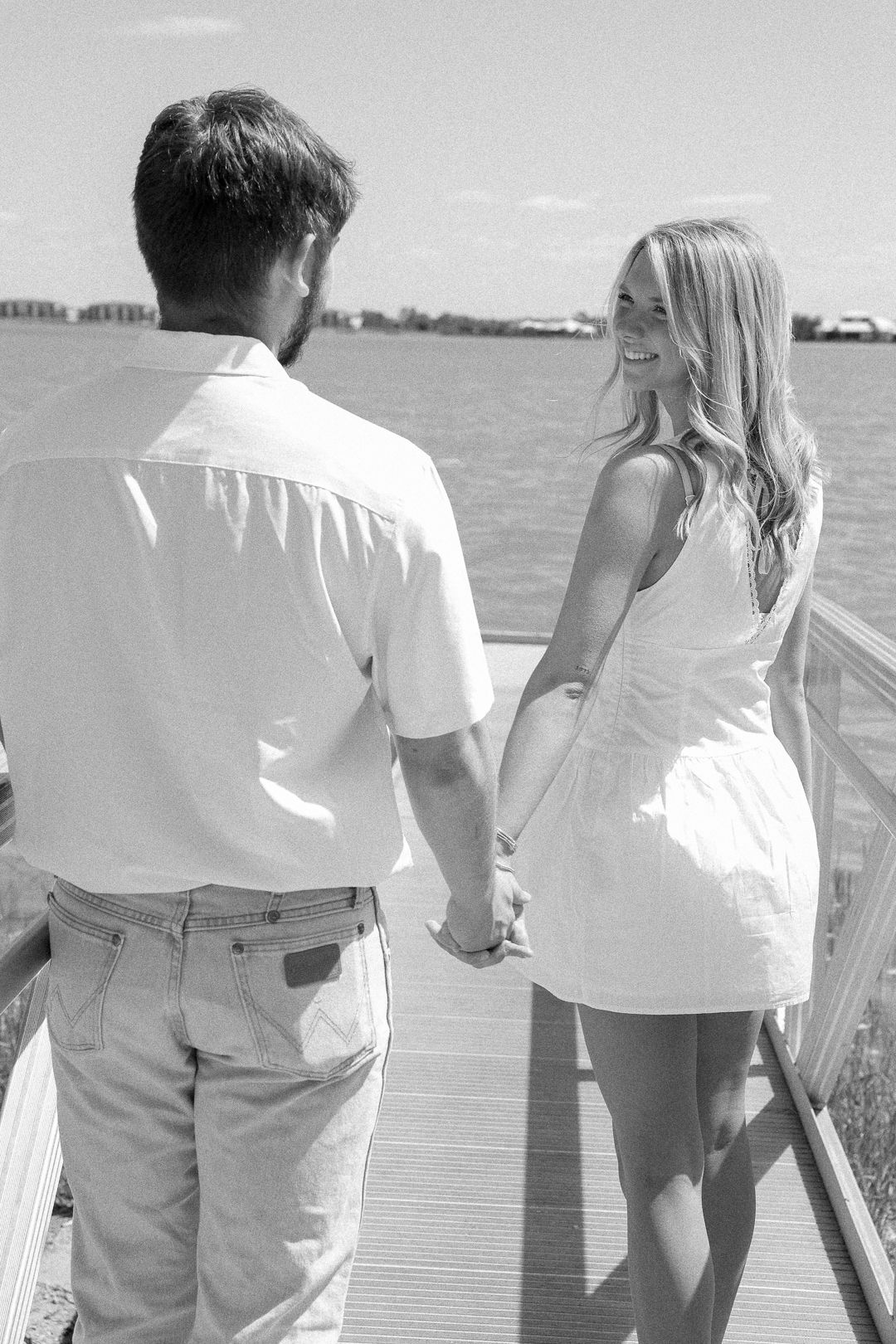 Black and white photo of a couple holding hands on a waterfront dock, woman smiling back.