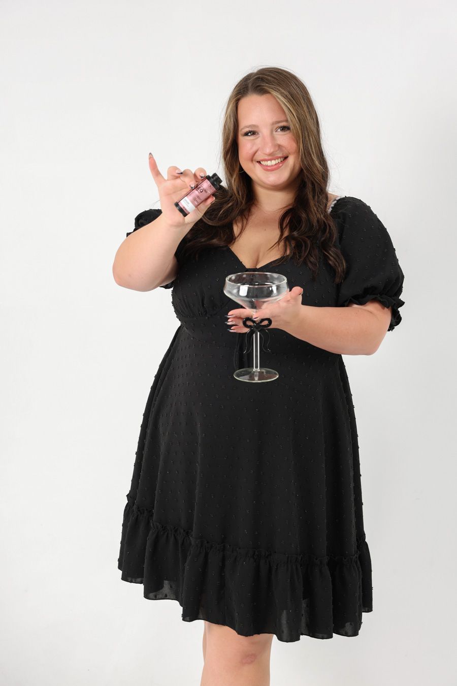 Woman in black dress smiling while holding a small bottle and coupe glass with black bow.