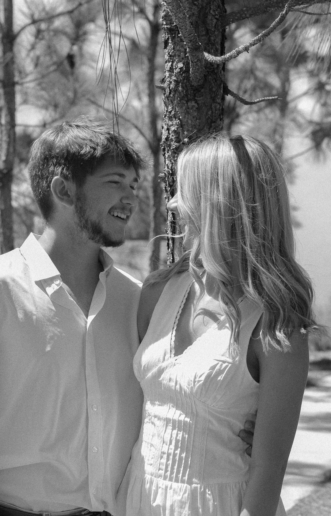 Black and white photo of a smiling couple standing by a tree in a sunny park.