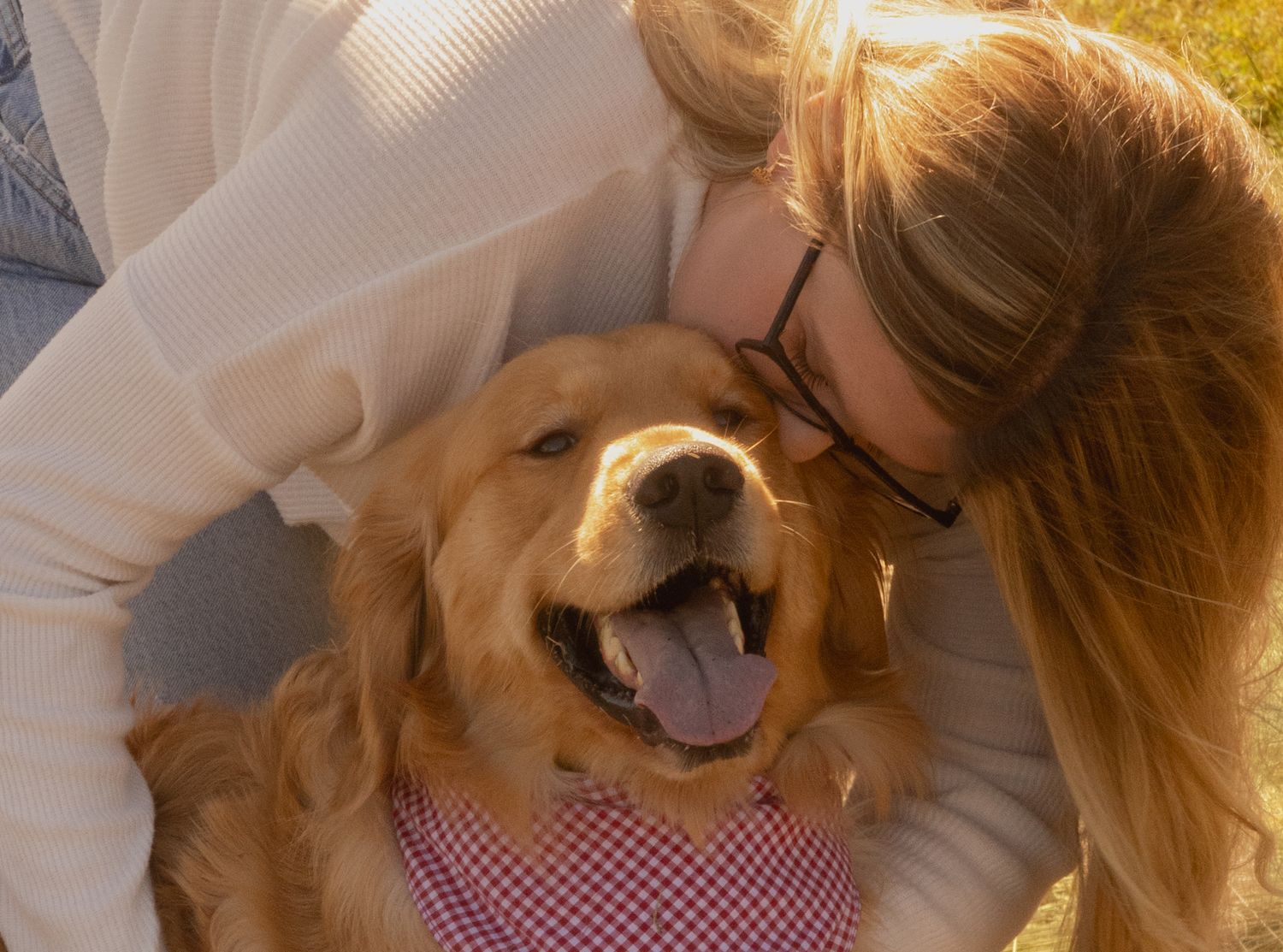 A happy golden retriever smiles with tongue out as a blonde woman in glasses hugs and kisses the dog outdoors.