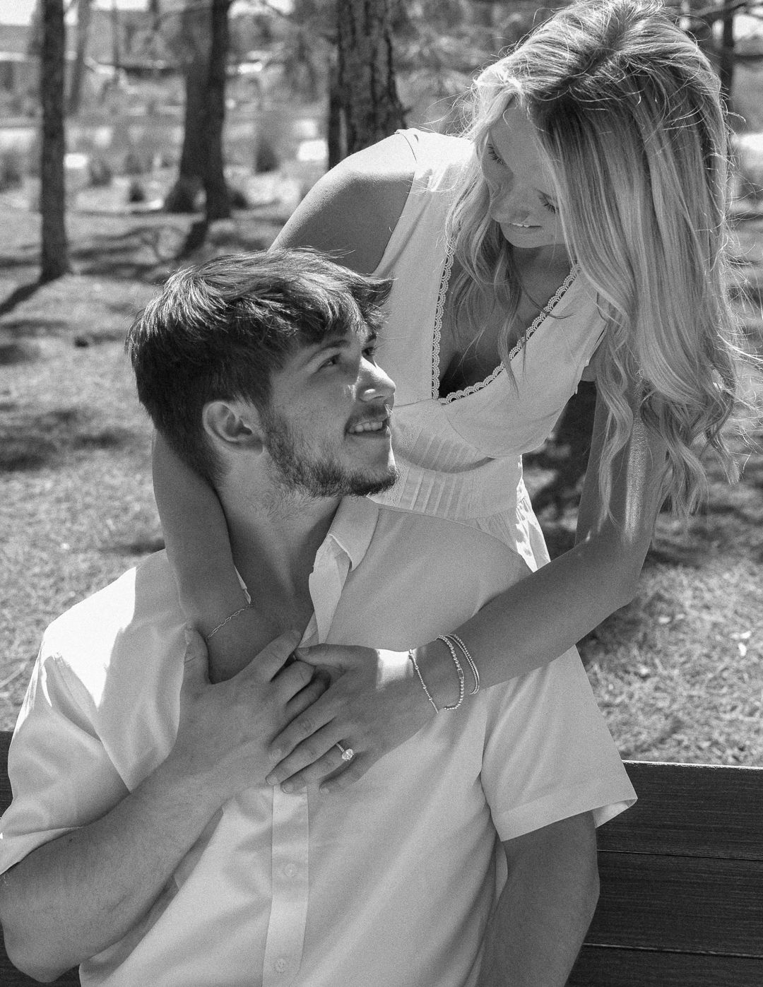 Black and white photo of a woman embracing a seated man from behind on a bench outdoors.