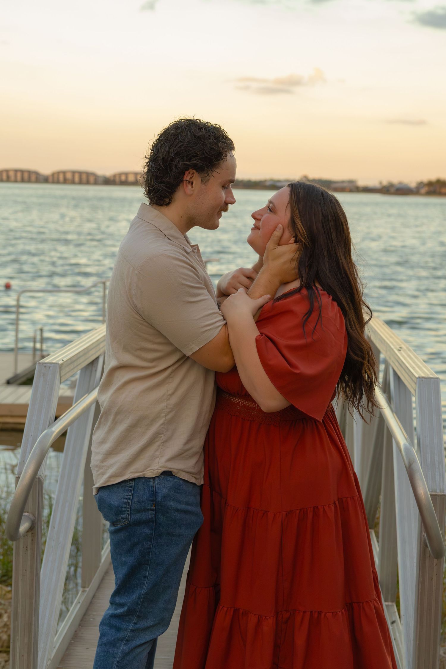 Couple embracing on a waterfront dock at sunset, man in tan shirt and jeans, woman in red dress.