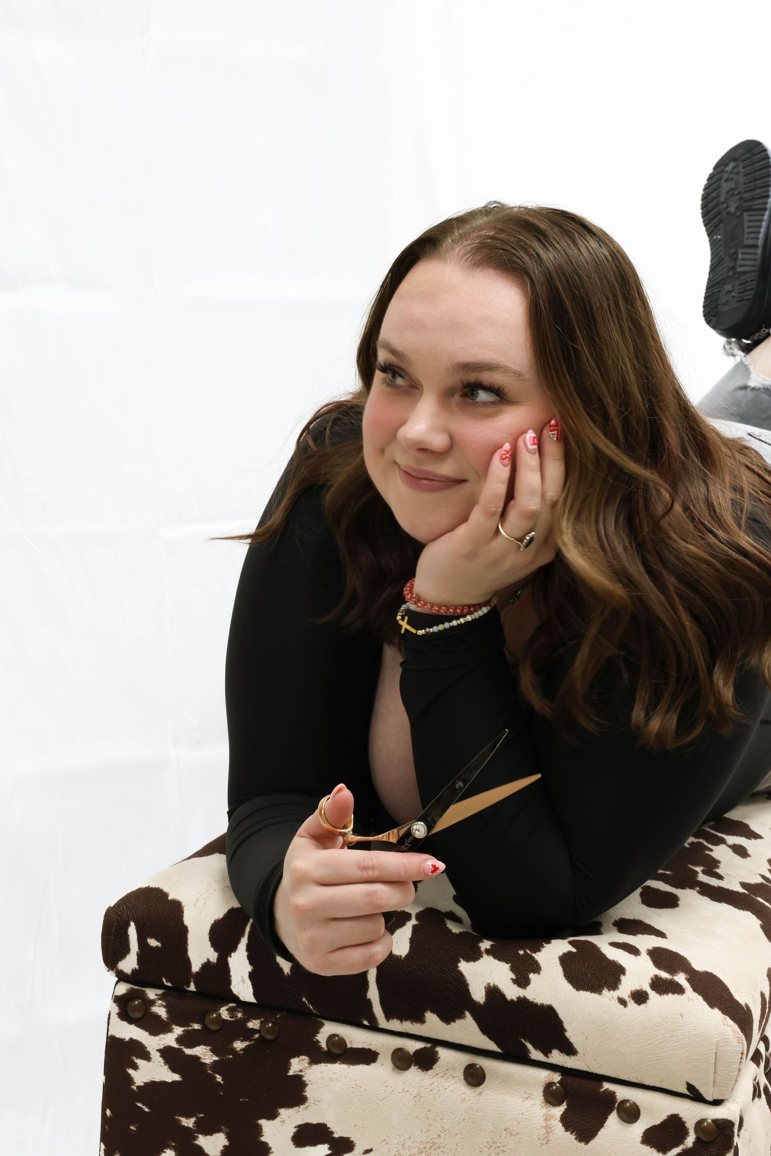 Young woman with wavy brown hair resting chin on hand, lying on cow print furniture, smiling thoughtfully.