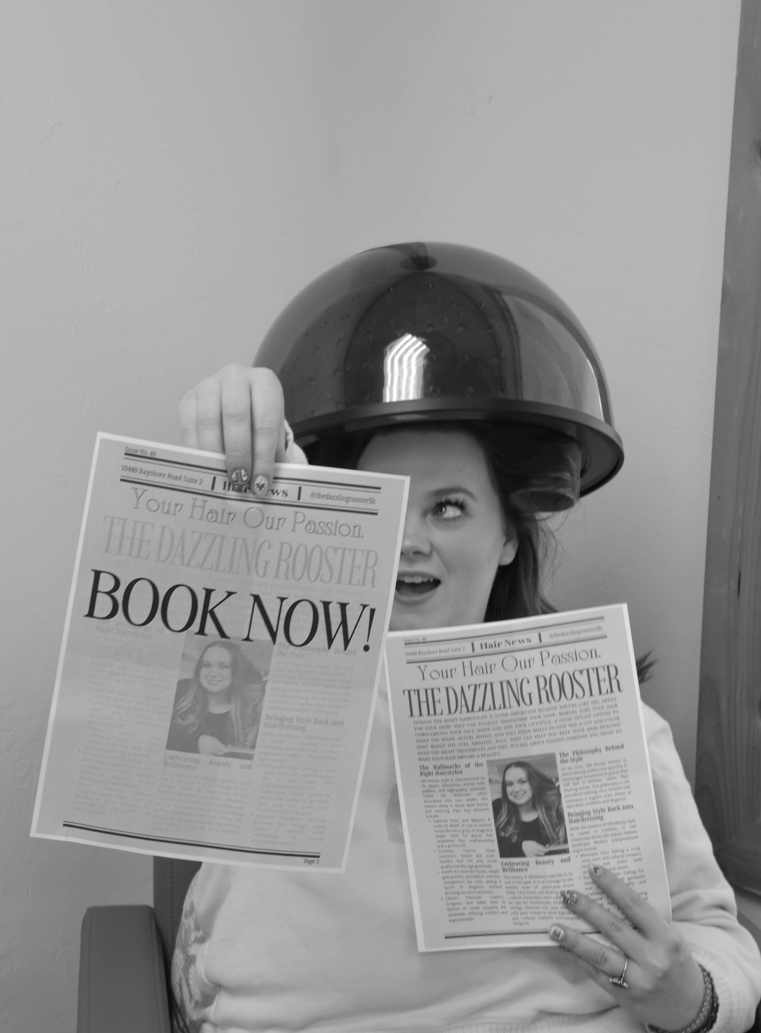Woman under salon hair dryer hood holding newspapers reading The Dazzling Booster Book Now in black and white.