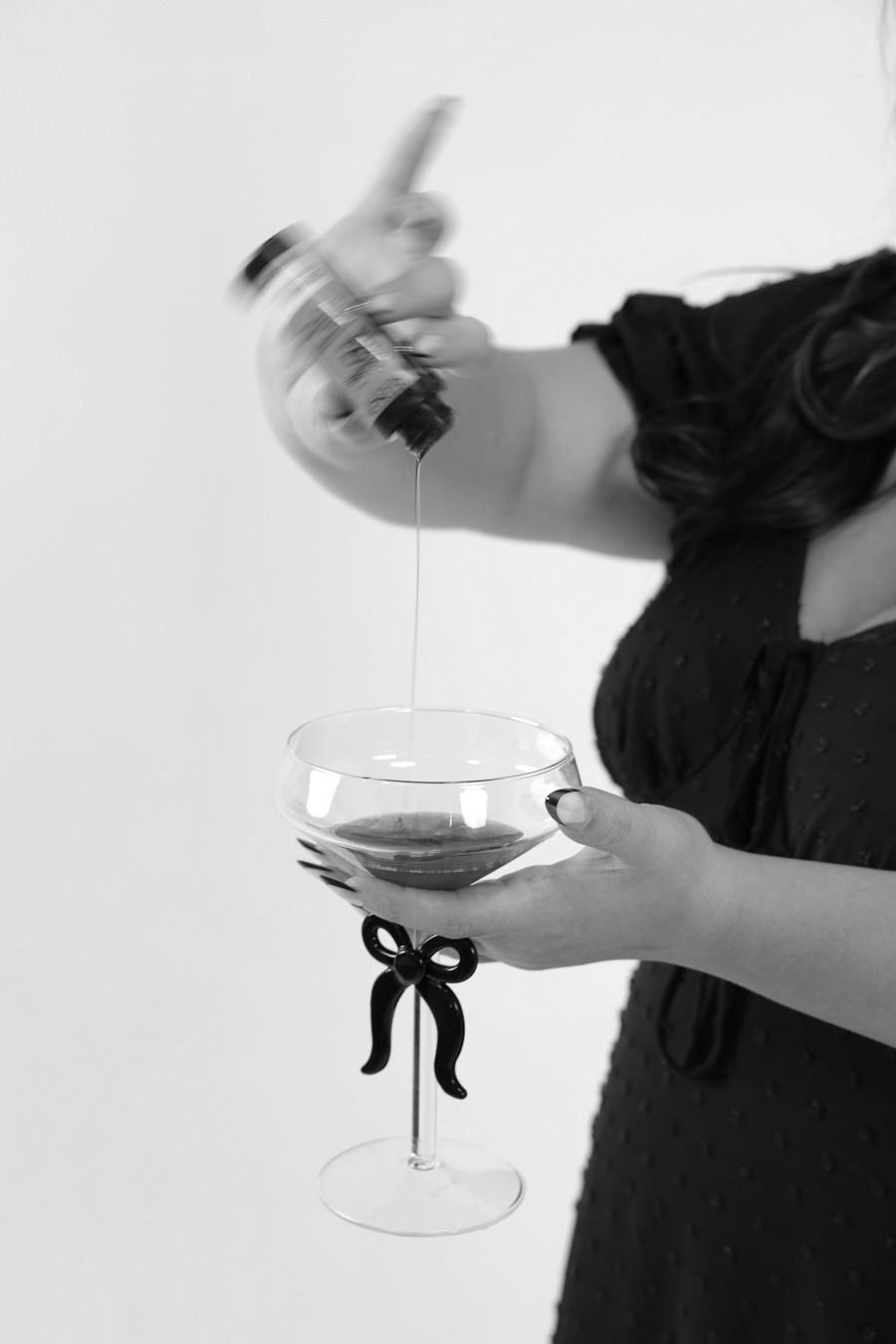 Black and white close-up of liquid being poured from a bottle into a bow-decorated coupe glass.