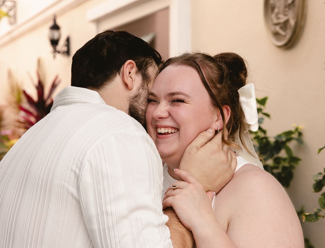 Bride laughing joyfully as groom kisses her cheek during wedding celebration outdoors.