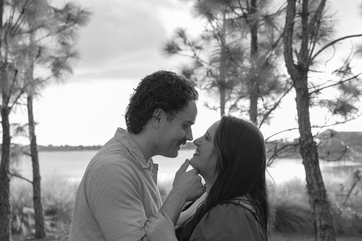 Black and white photo of couple nearly kissing among trees near a lake.