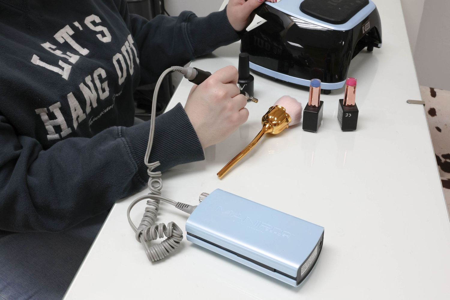 Person in Let's Hang Out sweatshirt using electric nail drill at white table with beauty tools.