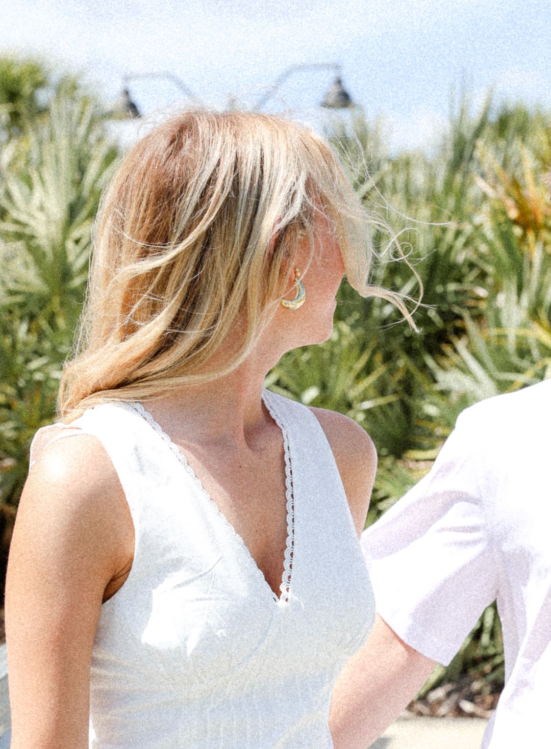 A blonde woman in a white dress with turquoise earrings standing outdoors on a sunny day.