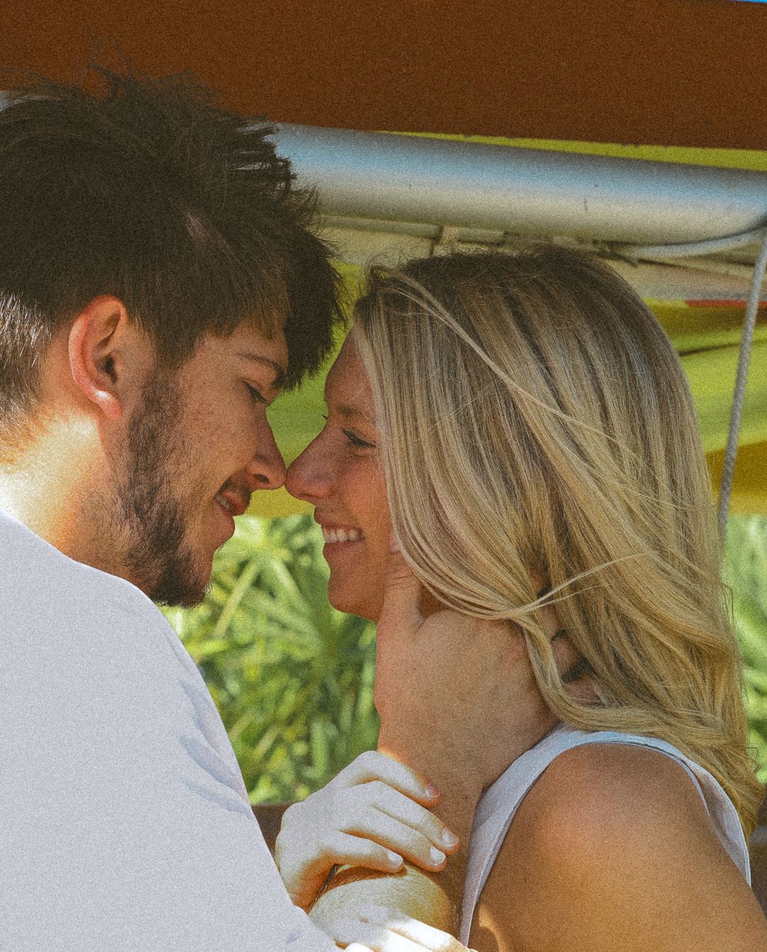 A couple shares a romantic moment, touching foreheads and smiling inside a vintage vehicle surrounded by greenery.