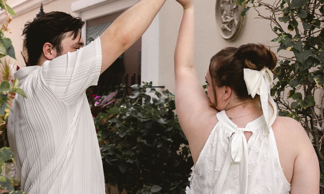 Couple in white outfits dancing outdoors, woman wearing backless halter top with bow, surrounded by lush greenery.