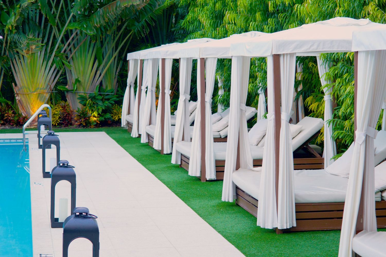 Luxury poolside cabanas with white curtains and lounge chairs beside a blue pool surrounded by tropical greenery.