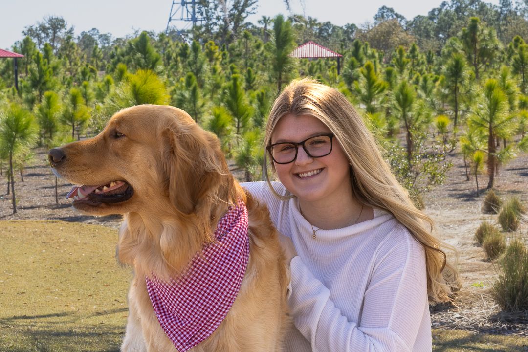 Young blonde girl with glasses hugging her Golden Retriever dog in a tree-lined park.