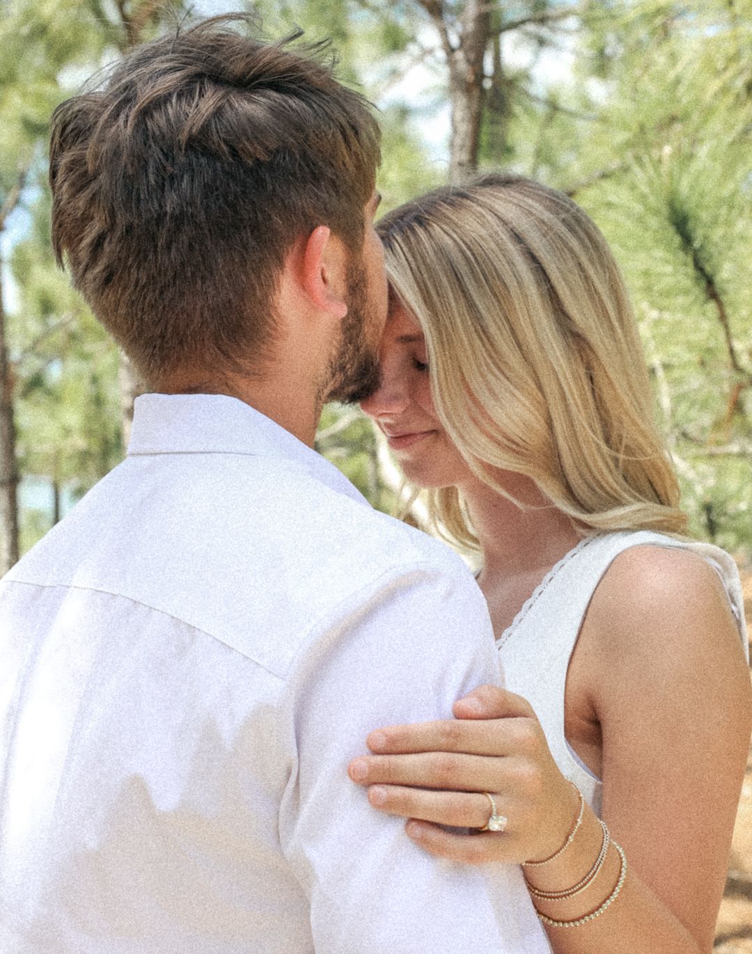 A couple in white clothing embraces closely among pine trees during an engagement photo session.