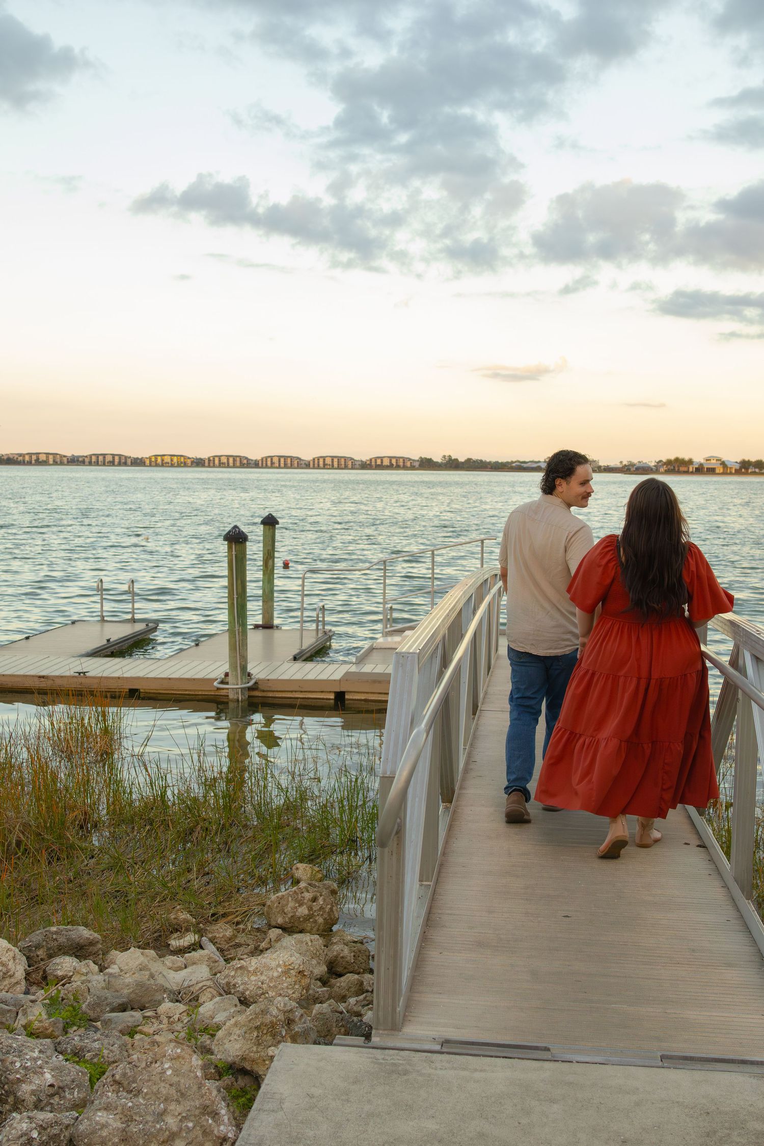Couple walks along a wooden dock toward the water at a peaceful sunset.