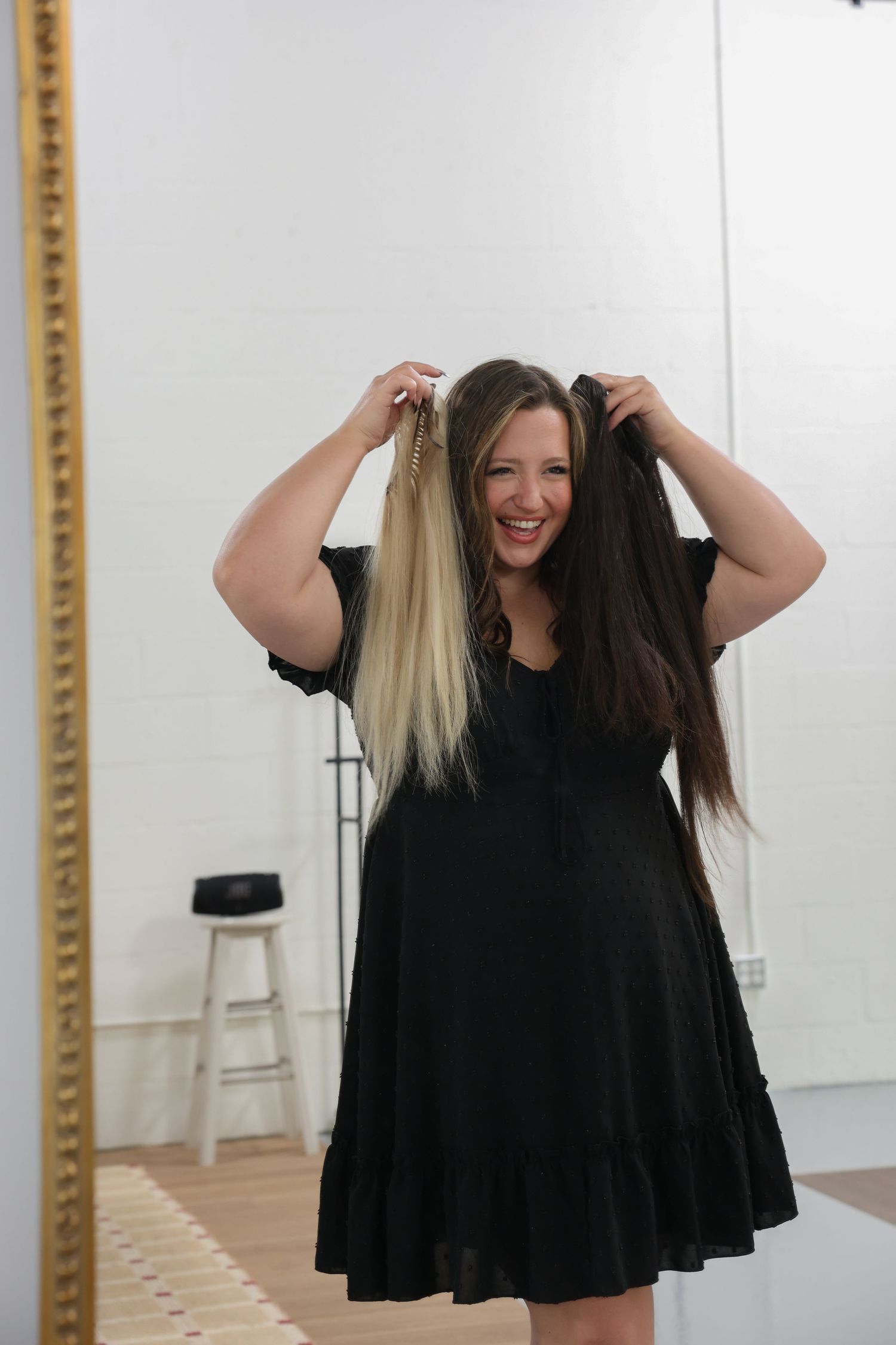 Smiling woman in black dress holding blonde and dark hair extensions in front of a gold mirror.
