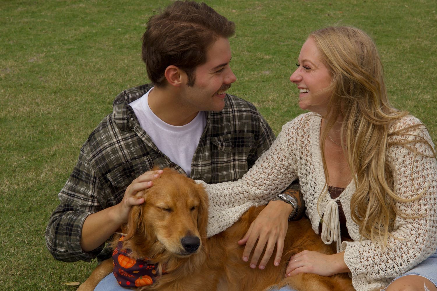 Smiling couple petting golden retriever wearing bandana while sitting on grass.