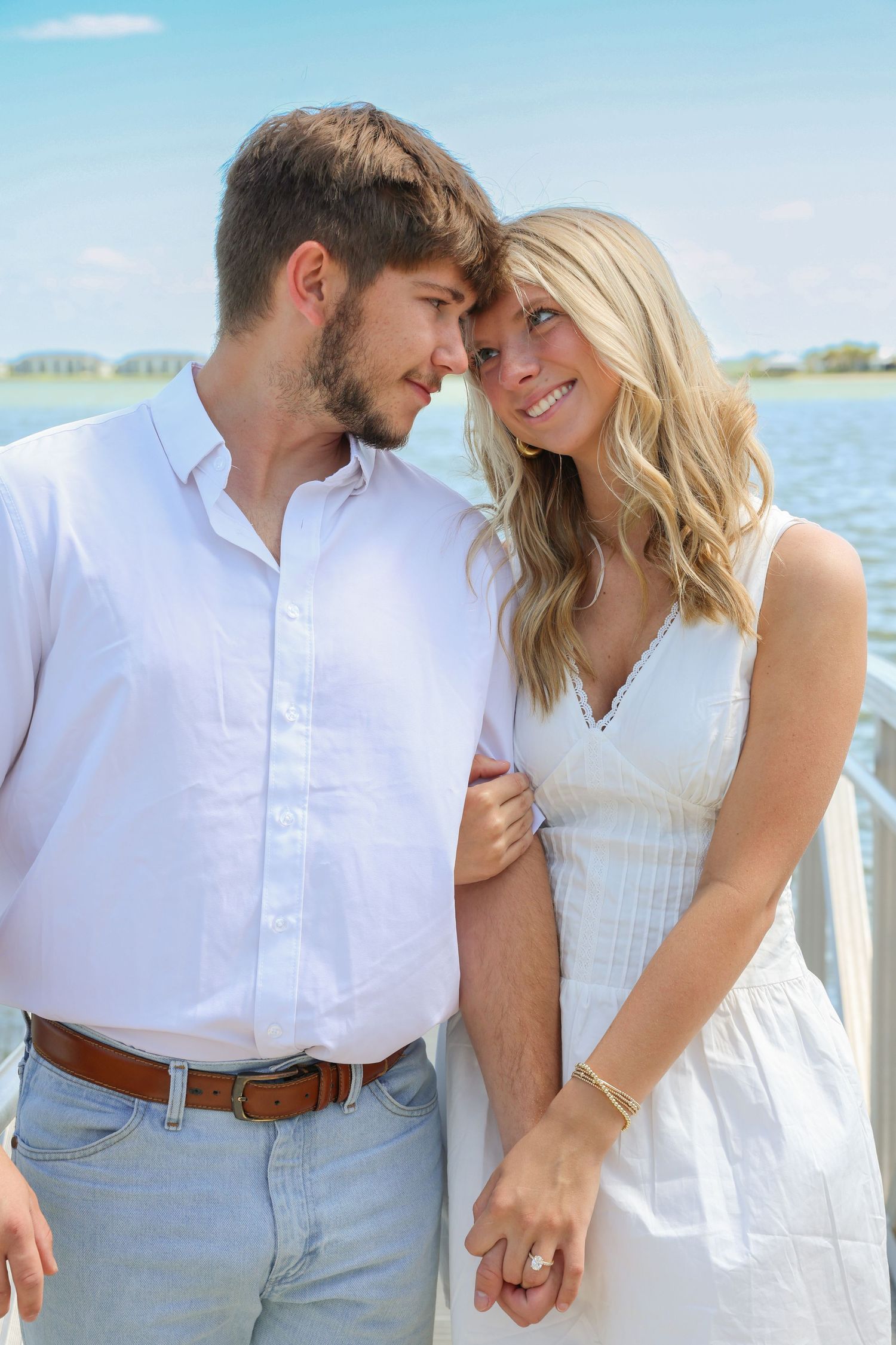 Couple in white outfits sharing a romantic moment by the water on a sunny day.